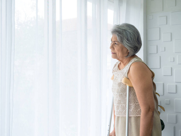 Asian senior woman white hair standing with walking crutches near curtain looking out glass window with copy space. Elderly lady patient using crutches walker. Strong health, medical care concepts. - Fotó, kép