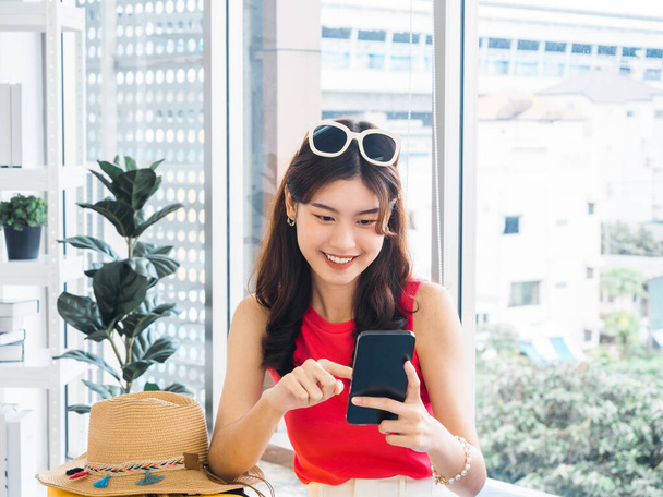 Young beautiful smiling Asian woman holding and using smart mobile phone, looking at screen. Happy Asian female wearing pink sleeveless, sunglasses, searching and planning for travel summer trip. - Foto, Imagen