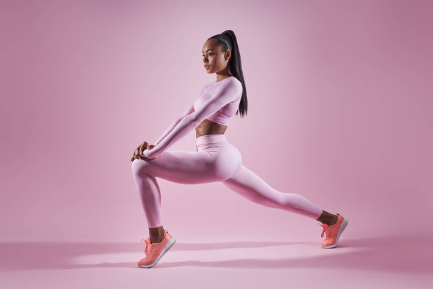 Beautiful young woman in sports clothing doing stretching exercises against pink background - Foto, afbeelding