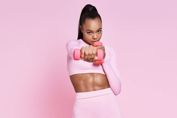 Concentrated young woman exercising with dumbbells while standing against pink background - Fotografie, Obrázek