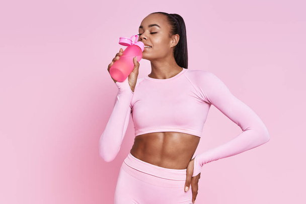 Happy African woman in sports clothing drinking water against pink background - Foto, Imagen