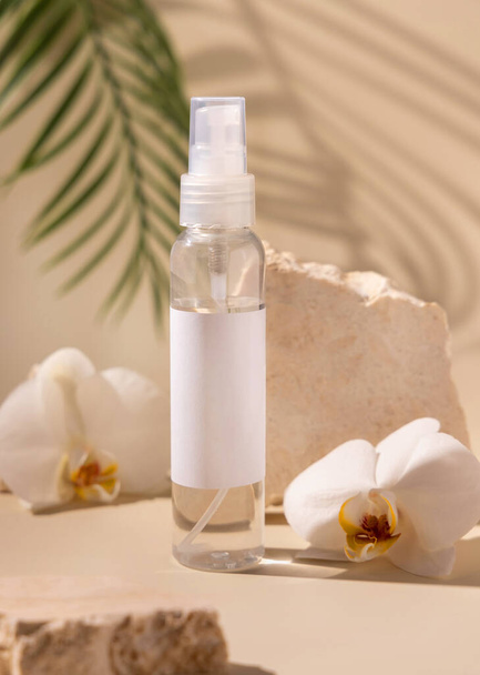 Spray dispencer bottle near white orchid flowers and beige stones on light beige close up, palm leaves hard shadows, mockup. Skincare beauty product package. Natural cosmetics, minimal compositio - Фото, изображение