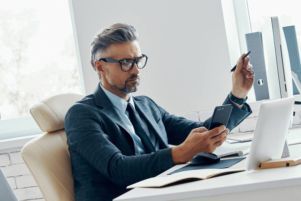 Уверенный бизнесмен, держащий смартфон и указывающий на монитор компьютера, сидя в офисе - Фото, изображение