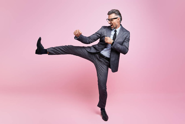 Mature man in formalwear throwing leg kick while standing against pink background - Photo, Image