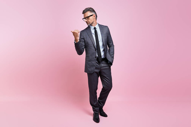 Confident mature man in full suit pointing away while standing against pink background - Foto, afbeelding