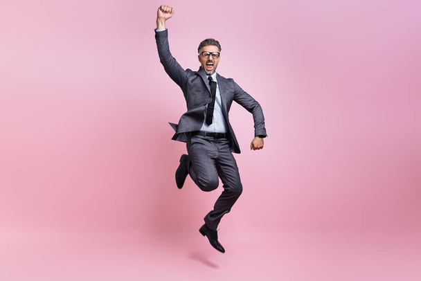 Excited mature man in full suit jumping and gesturing against pink background - Fotó, kép