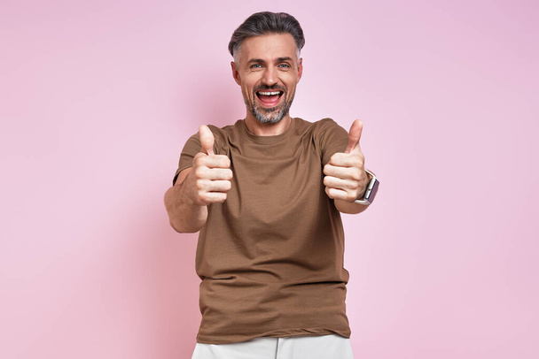 Happy mature man looking at camera and showing his thumbs up against pink background - Photo, Image