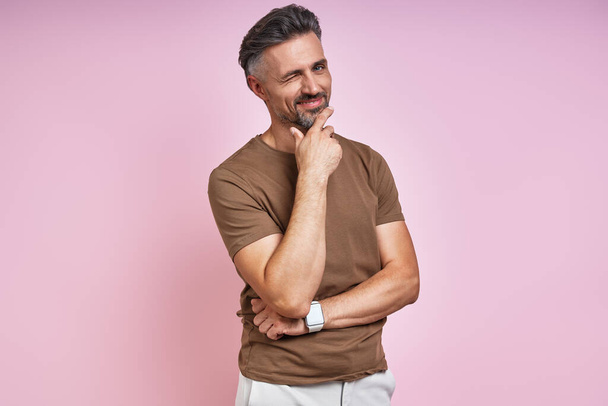 Handsome mature man holding hand on chin and winking while standing against pink background - Foto, immagini