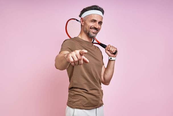 Happy mature man carrying tennis racket and pointing camera while standing against pink background - Foto, immagini