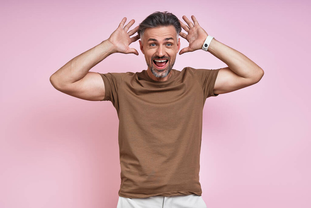 Surprised mature man looking at camera and gesturing while standing against pink background - Фото, изображение
