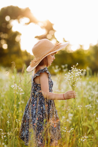 smiling child girl in a big mommys straw hat with bouquet of wildflowers in a green grassy meadow on summer sunny day. Happy childhood concept. Copy space - Photo, Image