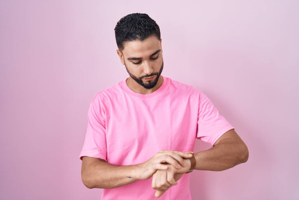 Латиноамериканец, стоящий на розовом фоне, проверяя время на наручных часах, расслабленный и уверенный в себе  - Фото, изображение
