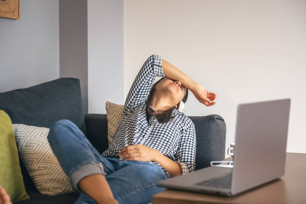 Κουρασμένος από τη δουλειά, μια νεαρή γυναίκα με ένα φορητό υπολογιστή βρίσκεται στον καναπέ. - Φωτογραφία, εικόνα