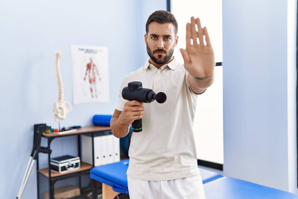 Pohledný hispánský muž držící terapeutickou masážní pistoli ve fyzioterapeutickém centru s otevřenou rukou dělá stopku s vážným a sebevědomým výrazem, obranné gesto  - Fotografie, Obrázek