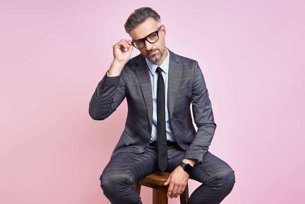 Handsome mature man in formalwear adjusting eyeglasses while sitting against pink background - Φωτογραφία, εικόνα