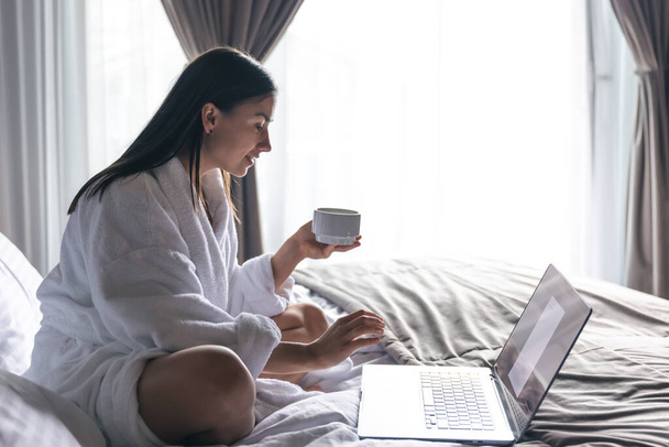 A young woman in a white coat is working on a laptop in bed with a cup of coffee. - Valokuva, kuva