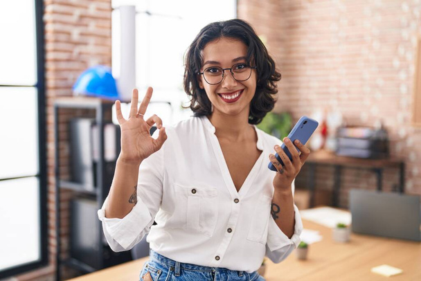 Young hispanic woman working at the office using smartphone doing ok sign with fingers, smiling friendly gesturing excellent symbol  - Fotoğraf, Görsel