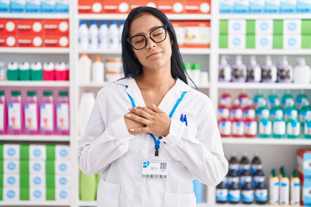 Mujer hispana trabajando en farmacia sonriendo con las manos en el pecho con los ojos cerrados y gesto agradecido en la cara. concepto de salud.  - Foto, Imagen