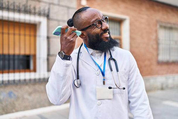 Giovane uomo afroamericano indossando uniforme medico ascolto messaggio audio dallo smartphone in ospedale - Foto, immagini