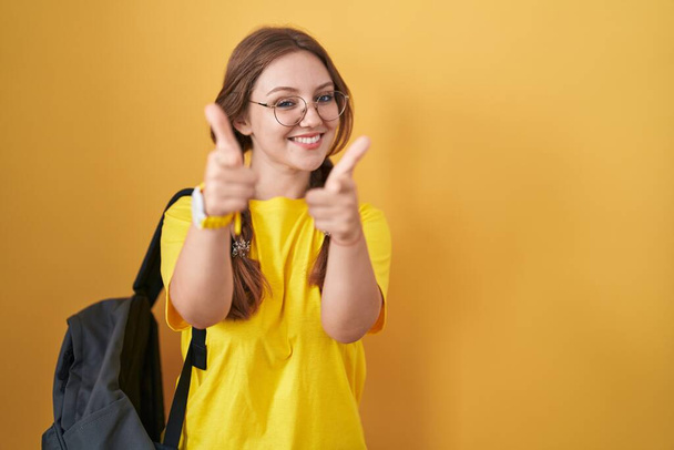 Νεαρή καυκάσια γυναίκα που φοράει φοιτητικό σακίδιο πάνω από κίτρινο φόντο δείχνοντας τα δάχτυλα της κάμερας με χαρούμενο και αστείο πρόσωπο. καλή ενέργεια και δονήσεις.  - Φωτογραφία, εικόνα