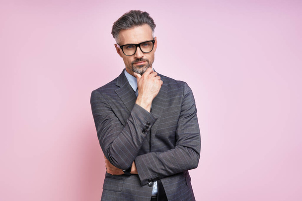 Handsome mature man in formalwear holding hand on chin while standing against pink background - Photo, Image