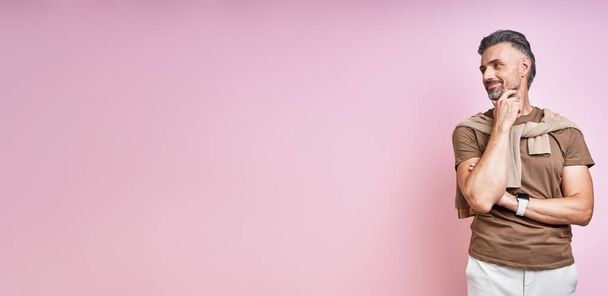 Handsome mature man holding hand on chin and looking away while standing against pink background - Foto, afbeelding