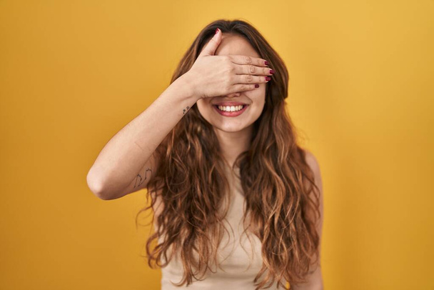 Fiatal spanyol nő áll a sárga háttér felett mosolyogva és nevetve kéz a kézben eltakarja a szemét meglepetés. vak fogalom.  - Fotó, kép