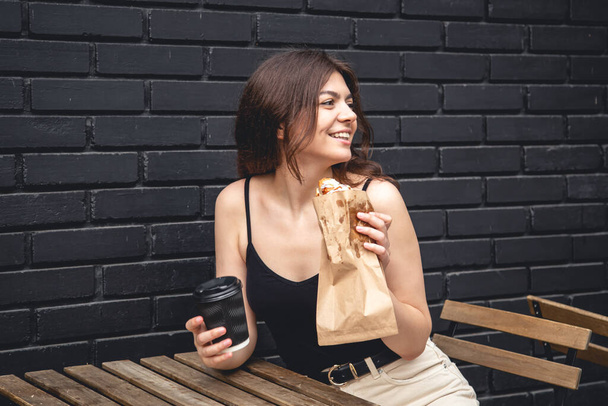 A young woman with a croissant and a cup of coffee on the background of a black brick wall of the cafe exterior. - Foto, Imagem