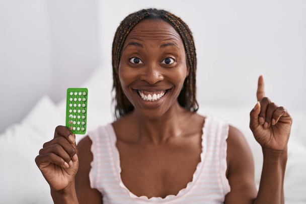 African american woman holding birth control pills surprised with an idea or question pointing finger with happy face, number one  - Foto, Bild