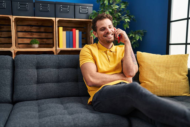 Young hispanic man talking on the smartphone sitting on sofa at home - Foto, Imagem