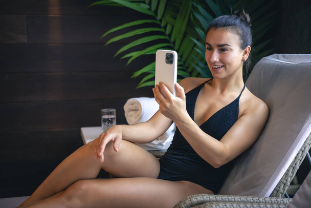 A young woman in a swimsuit is relaxing in a spa complex and using a smartphone. - Fotografie, Obrázek