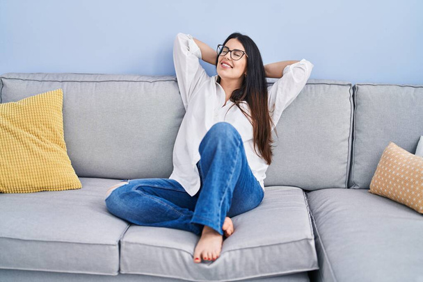 Молода іспаномовна жінка розслабилася руками на голові, сидячи на дивані вдома
 - Фото, зображення