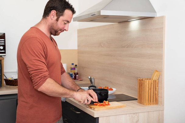 Ο άνθρωπος κόβει μια ντομάτα με ένα μαύρο μαχαίρι σε μια ξύλινη σανίδα κοπής στην κουζίνα του. - Φωτογραφία, εικόνα