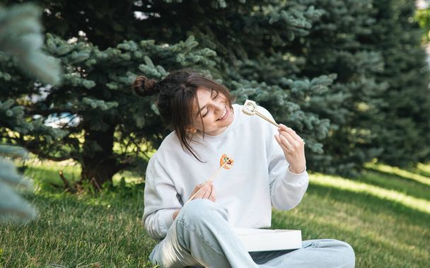 Fröhliche junge Frau, die im Gras sitzt und Sushi im Park isst, Picknick in der Natur, Sushi-Lieferkonzept. - Foto, Bild