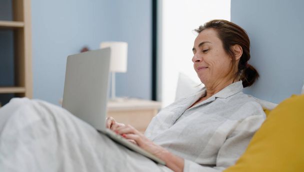 Middelbare leeftijd Spaanse vrouw met behulp van laptop zittend op bed in de slaapkamer - Foto, afbeelding
