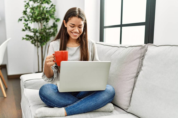 Молодая латиноамериканка с ноутбуком и пьет кофе сидя дома на диване - Фото, изображение