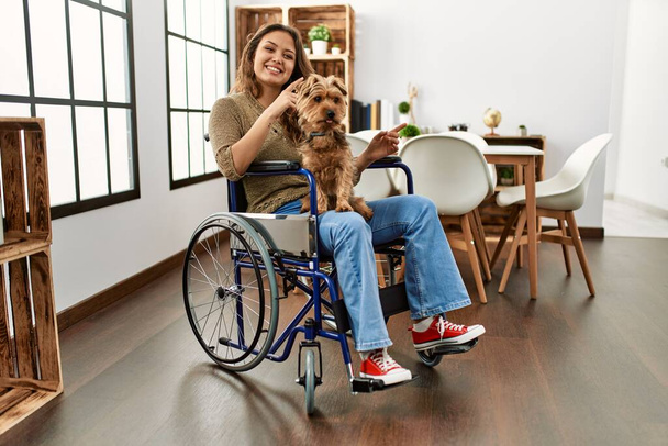 Młoda Latynoska dziewczyna siedząca na wózku inwalidzkim w domu uśmiechnięta, radosna i wskazująca dłonią na aparat fotograficzny.  - Zdjęcie, obraz
