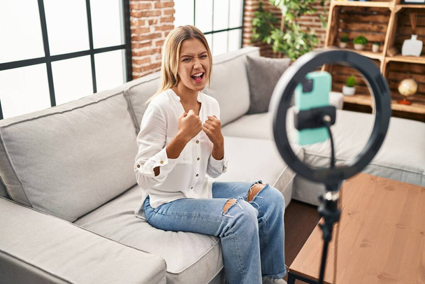 Young blonde woman recording vlog tutorial with smartphone at home celebrating surprised and amazed for success with arms raised and eyes closed  - Foto, Bild