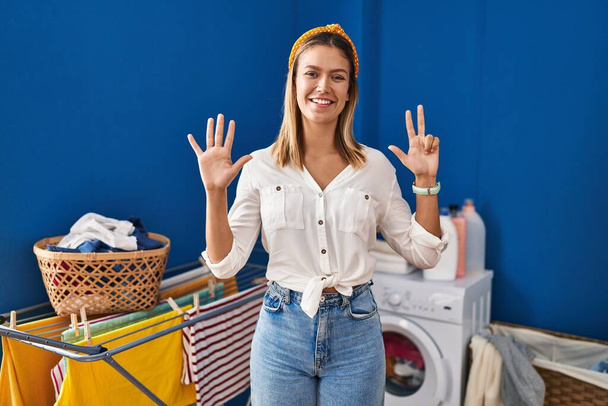 Νεαρή ξανθιά γυναίκα στο πλυσταριό δείχνει και δείχνει προς τα πάνω με τα δάχτυλα νούμερο οκτώ, ενώ χαμογελά αυτοπεποίθηση και χαρούμενος.  - Φωτογραφία, εικόνα