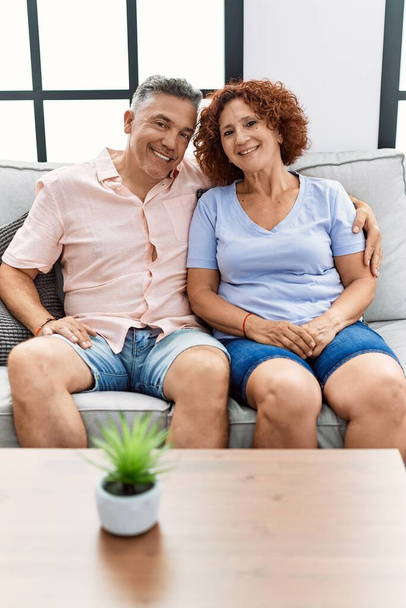 mężczyzna i kobieta w średnim wieku para uśmiecha się pewnie przytulając się siedząc na kanapie w domu - Zdjęcie, obraz