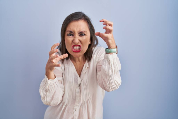 Middle age hispanic woman standing over blue background shouting frustrated with rage, hands trying to strangle, yelling mad  - Fotó, kép