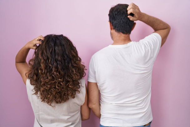 Middle age hispanic couple together over pink background backwards thinking about doubt with hand on head  - Φωτογραφία, εικόνα