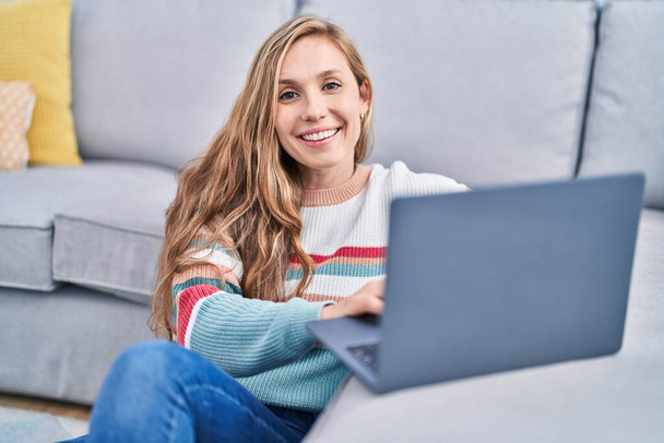 Junge blonde Frau sitzt mit Laptop zu Hause auf dem Boden - Foto, Bild