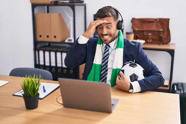 Schöner hispanischer Mann schaut Fußballspiel im Büro gestresst und frustriert mit Hand auf Kopf, überraschtem und wütendem Gesicht  - Foto, Bild