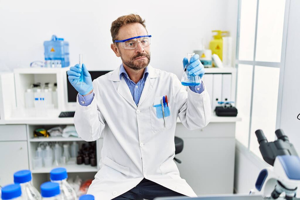 Чоловік середнього віку, працюючи в науковій лабораторії, тримає хімічні продукти, посміхаючись, дивлячись в бік і дивлячись на мислення
.  - Фото, зображення