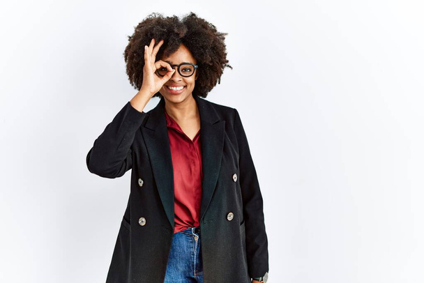 Femme afro-américaine avec des cheveux afro portant une veste d'affaires et des lunettes faisant un geste correct avec un sourire à la main, un œil regardant à travers les doigts avec un visage heureux.  - Photo, image