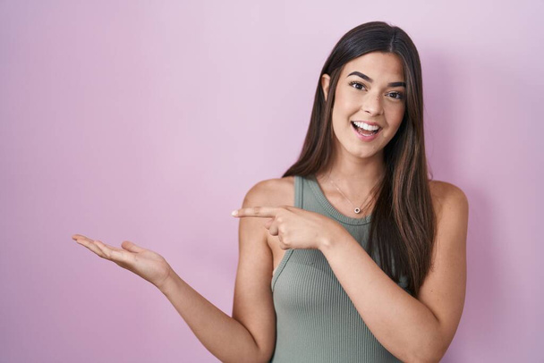 Latino nainen seisoo vaaleanpunainen tausta hämmästynyt ja hymyillen kameralle samalla esittää käsi ja osoittaa sormella.  - Valokuva, kuva