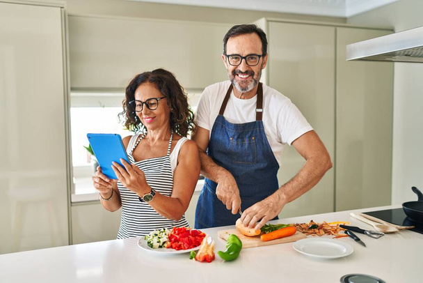 Latynoska para w średnim wieku uśmiecha się pewnie gotując i używając touchpada w kuchni - Zdjęcie, obraz
