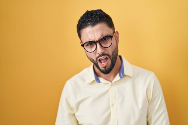 Hispanic young man wearing business clothes and glasses in shock face, looking skeptical and sarcastic, surprised with open mouth  - Photo, Image