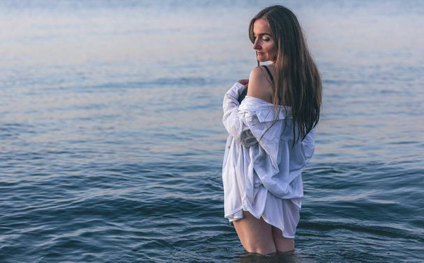 A woman in a bathing suit and a white shirt in the sea at sunset, copy space. - Fotografie, Obrázek